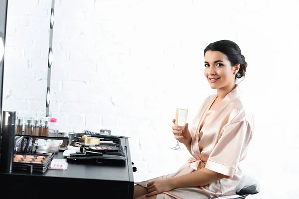 Vista lateral da noiva sorridente em roupão de banho de seda e véu com copo de champanhe sentado à mesa com cosméticos — Fotografia de Stock