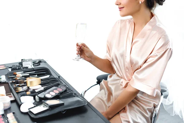 Partial view of smiling bride in silk bathrobe and veil with glass of champagne sitting at tabletop with cosmetics — Stock Photo