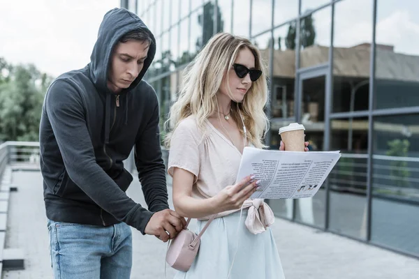 Mulher com café para ir ler jornal enquanto ladrão pickpocketing de sua bolsa — Fotografia de Stock