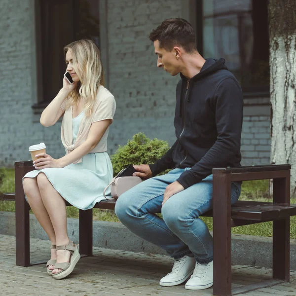 Man pickpocketing wallet from bag of woman with coffee talking on smartphone on bench in park — Stock Photo