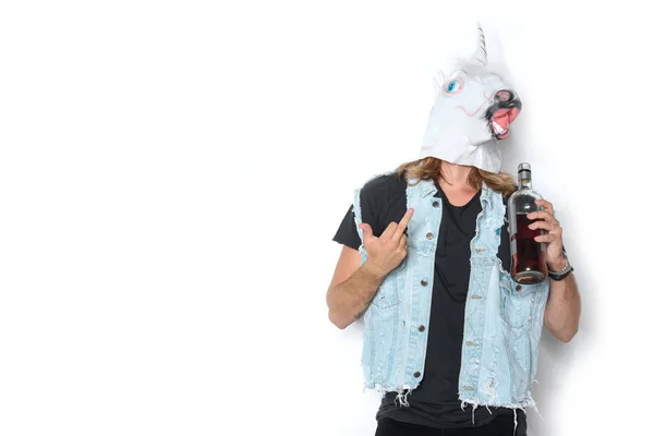 Mann in Einhorn-Maske und Jeansweste mit Flasche Rum zeigt Mittelfinger auf Weiß — Stockfoto