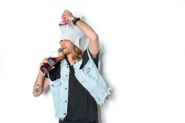 Young man in unicorn mask and denim vest lifting mask and drinking rum on white — Stock Photo