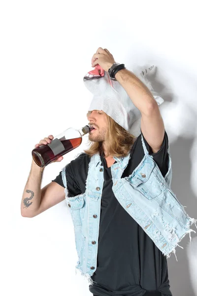 Man in unicorn mask and denim vest lifting mask and drinking rum on white — Stock Photo