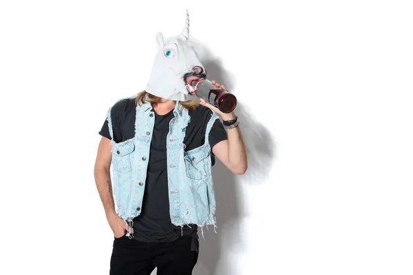 Homme en masque de licorne et gilet en denim boire du rhum à travers le masque sur blanc — Photo de stock