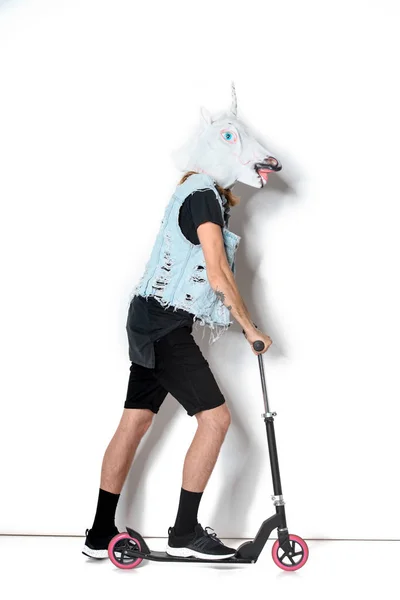 Vista laterale dell'uomo in maschera di unicorno e giubbotto di jeans equitazione scooter su bianco — Foto stock