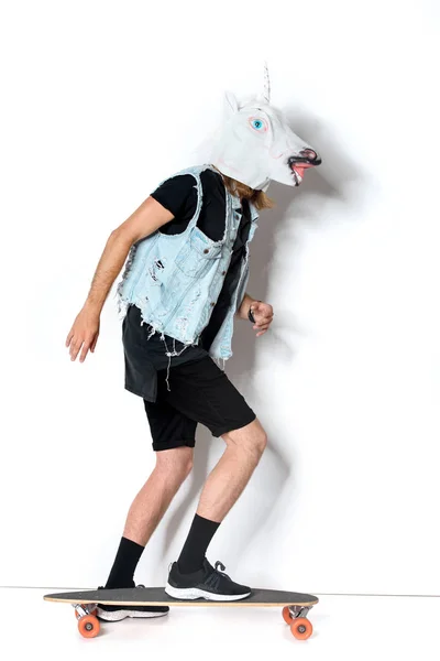 Vista lateral do homem em máscara de unicórnio e colete de ganga andar de skate no branco — Fotografia de Stock
