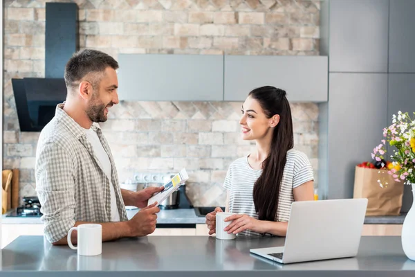 Seitenansicht eines lächelnden Paares mit Zeitung und Tasse Kaffee, das sich am Tresen mit Laptop in der Küche unterhält — Stockfoto