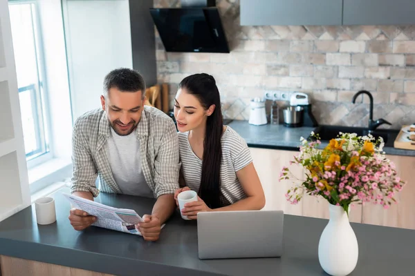 Portrait of smiling married couple reading newspaper together at counter with laptop in kitchen — Stock Photo