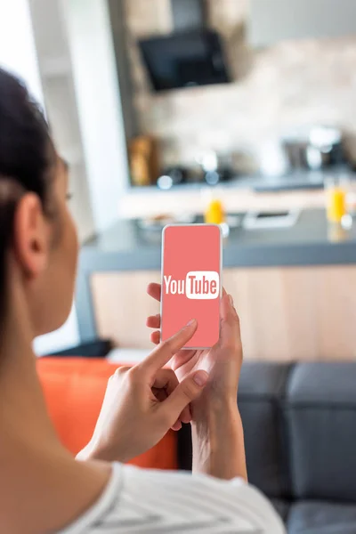 Enfoque selectivo de la mujer utilizando el teléfono inteligente con logotipo de youtube en la pantalla en la cocina - foto de stock