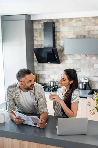 Retrato de pareja casada con periódico y taza de café de pie en el mostrador con ordenador portátil en la cocina - foto de stock