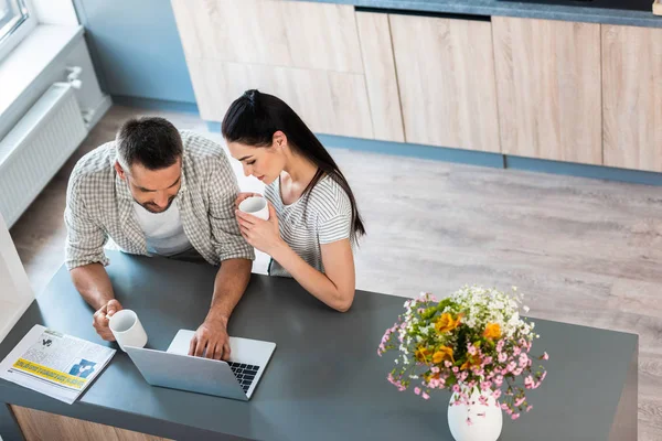 Vista de ángulo alto de la pareja casada con tazas de café utilizando el ordenador portátil juntos en el mostrador en la cocina - foto de stock