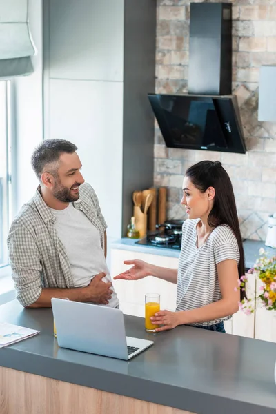 Vista lateral de pareja con vasos de jugo conversando en mostrador con laptop en cocina — Stock Photo