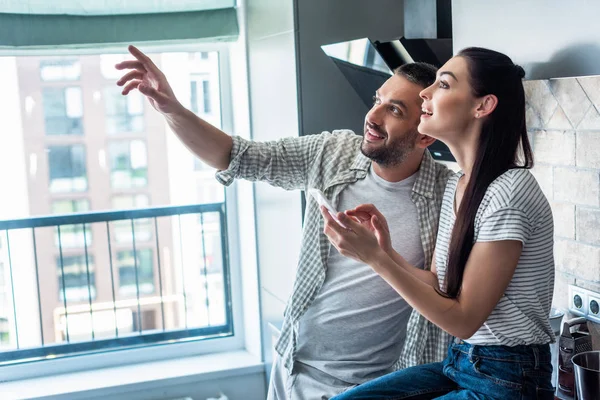 Вид збоку усміхненої пари зі смартфоном, який дивиться разом на кухню, концепція розумного будинку — стокове фото