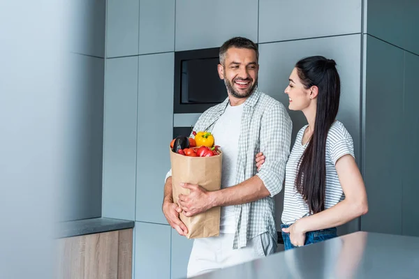 Casal alegre com pacote de papel cheio de legumes frescos em pé na cozinha em casa — Fotografia de Stock