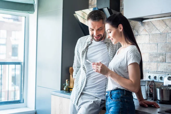 Couple souriant utilisant smartphone ensemble dans la cuisine, concept de maison intelligente — Photo de stock