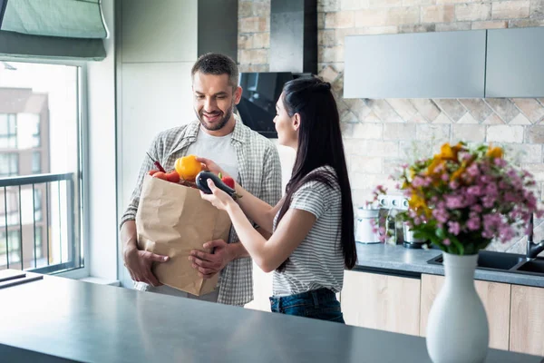 Casal com pacote de papel cheio de vegetais frescos para jantar na cozinha em casa — Fotografia de Stock