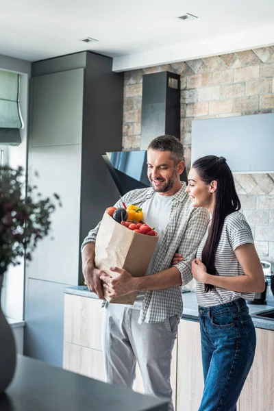 Happy married couple with paper package full of fresh vegetables for dinner in kitchen at home — Stock Photo