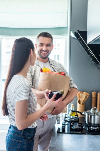 Feliz pareja casada con paquete de papel lleno de verduras frescas para la cena en la cocina en casa - foto de stock