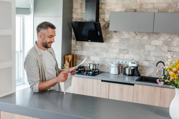 Vista lateral del hombre barbudo sonriente leyendo el periódico mientras está de pie en el mostrador en la cocina - foto de stock