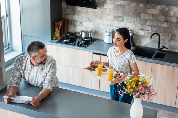 Uomo con giornale in piedi al bancone e guardando moglie con colazione su vassoio di legno in cucina — Foto stock