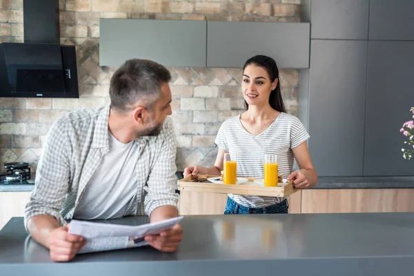Mann mit Zeitung steht am Tresen und schaut Frau mit Frühstück auf Holztablett in Küche an — Stockfoto