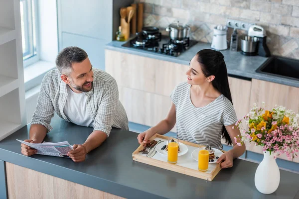 Hombre con periódico de pie en el mostrador y mirando a la esposa con el desayuno en bandeja de madera en la cocina - foto de stock