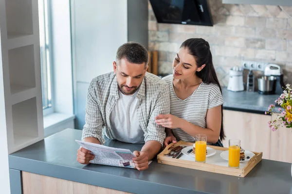 Мужчина читает газету с женой и завтраком рядом на деревянном подносе на кухне — стоковое фото