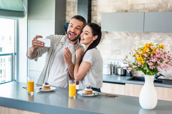 Sonriente pareja tomando selfie en teléfono inteligente en el mostrador con desayuno casero en la cocina - foto de stock