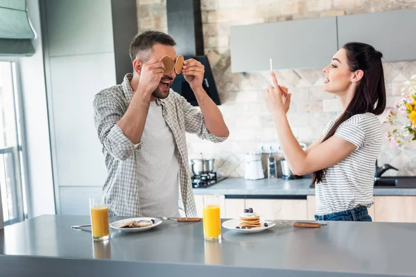 Mulher tirando foto de marido brincando com café da manhã no balcão na cozinha — Fotografia de Stock