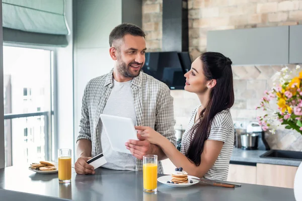 Casal sorrindo com tablet no balcão com café da manhã caseiro na cozinha — Fotografia de Stock