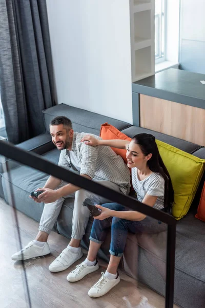 Happy married couple with gamepads playing video games together while sitting on sofa at home — Stock Photo