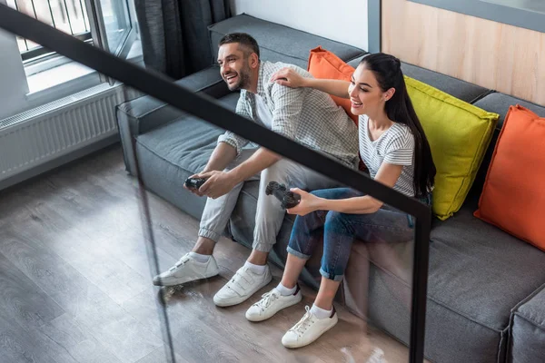 Married couple with gamepads playing video games together while sitting on sofa at home — Stock Photo