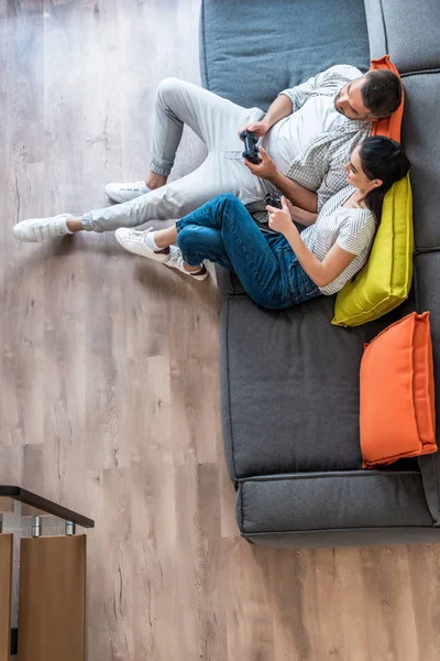 Vue aérienne du couple marié avec des manettes de jeu jouant ensemble à des jeux vidéo assis sur le canapé à la maison — Photo de stock