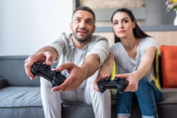Enfoque selectivo de la pareja casada con gamepads jugando videojuegos juntos mientras están sentados en el sofá en casa - foto de stock