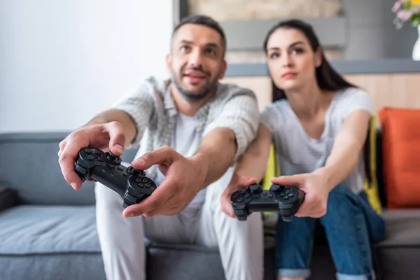 Foyer sélectif du couple marié avec des manettes de jeu jouer à des jeux vidéo ensemble tout en étant assis sur le canapé à la maison — Photo de stock