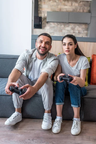 Couple marié avec des manettes de jeu jouer à des jeux vidéo ensemble tout en étant assis sur le canapé à la maison — Photo de stock