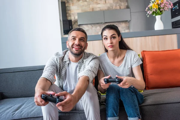 Retrato de pareja casada con gamepads jugando videojuegos juntos mientras están sentados en el sofá en casa - foto de stock