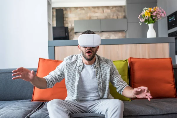 Homme émotionnel portant un casque de réalité virtuelle assis sur le canapé à la maison — Photo de stock