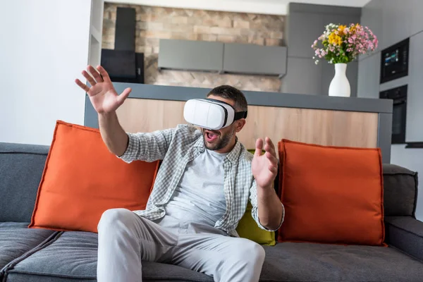 Homem emocional vestindo fone de ouvido realidade virtual enquanto sentado no sofá em casa — Fotografia de Stock