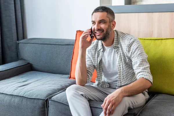 Sorrindo barbudo homem sentado no sofá e falando no smartphone em casa — Fotografia de Stock