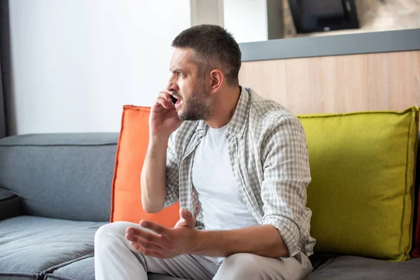 Эмоциональный бородатый мужчина сидит на диване и разговаривает на смартфоне дома — стоковое фото