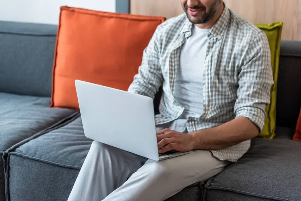 Vista parcial del hombre sonriente usando el ordenador portátil en el sofá en casa - foto de stock