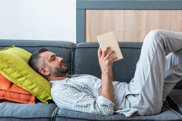 Vue latérale du livre de lecture homme barbu tout en étant couché sur le canapé à la maison — Photo de stock