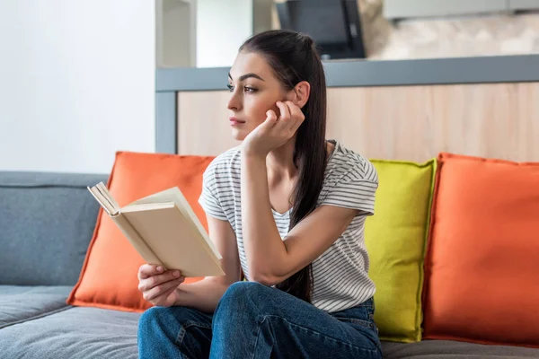 Retrato de mulher pensativa olhando para longe ao ler livro no sofá em casa — Fotografia de Stock