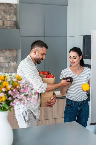 Sonriente pareja casada poniendo verduras frescas en la nevera en la cocina en casa - foto de stock