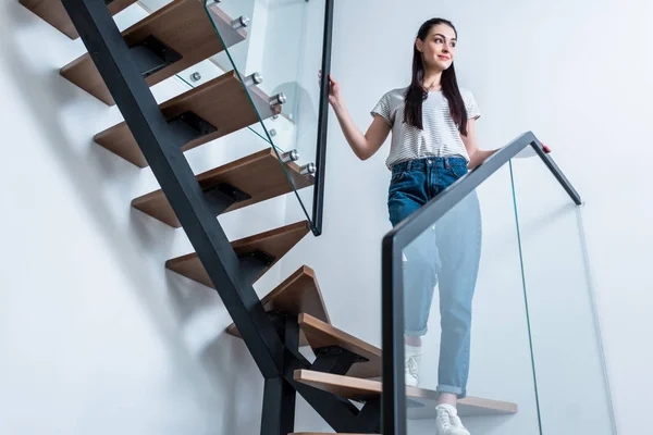 Baixo ângulo de visão de mulher sorridente em jeans olhando para longe, enquanto em pé nas escadas em casa — Fotografia de Stock