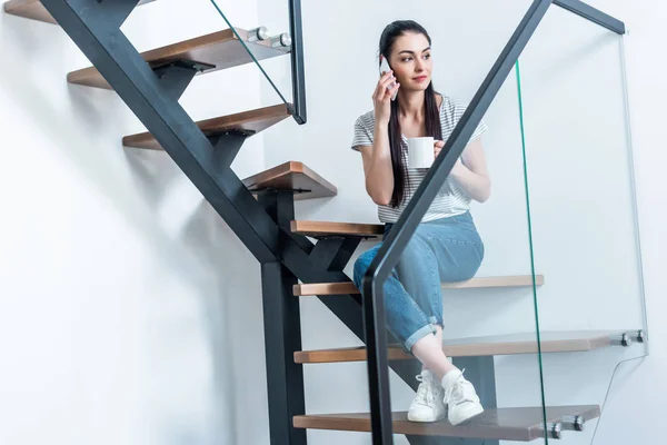 Frau mit Tasse Kaffee in der Hand spricht auf Smartphone, während sie zu Hause auf der Treppe sitzt — Stockfoto