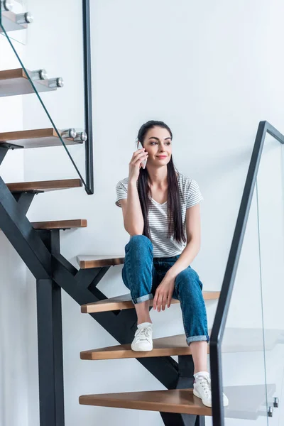Mujer sonriente sentada en las escaleras y hablando en el teléfono inteligente en casa - foto de stock