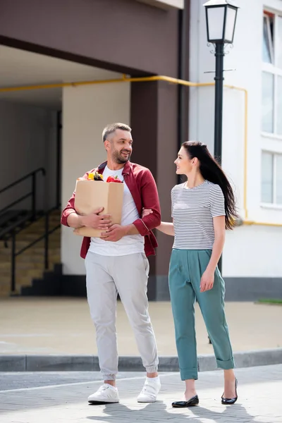 Couple souriant avec paquet de papier plein de légumes frais marchant dans la rue — Photo de stock