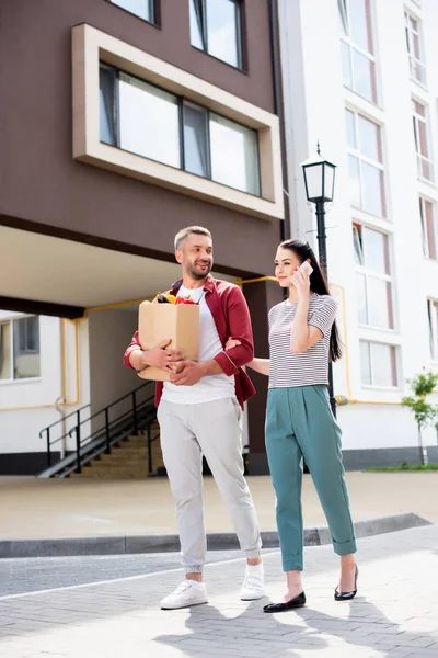 Woman talking on smartphone while walking together with husband after shopping on street — Stock Photo